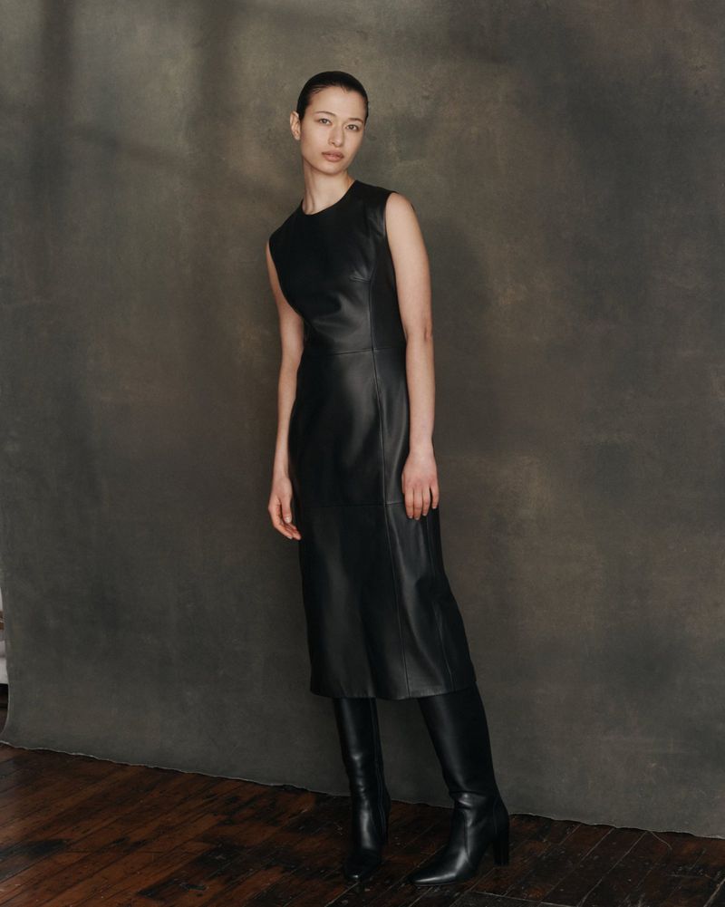 A full shot of a female fashion model in a sleeveless black dress and black leather boots, posing against a dark grey mottled backdrop.