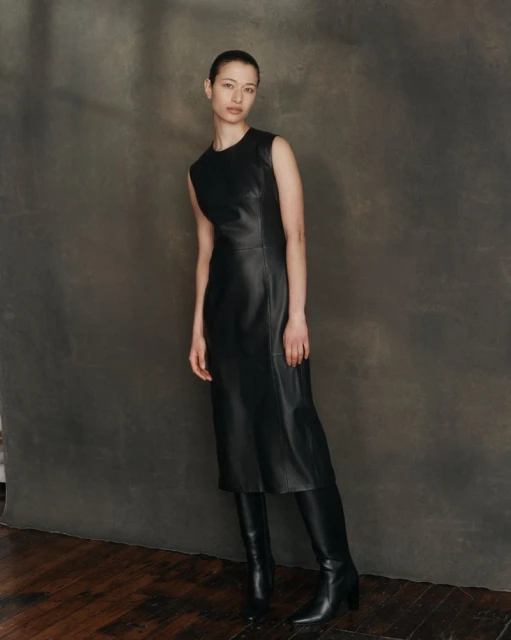 A full shot of a female fashion model in a sleeveless black dress and black leather boots, posing against a dark grey mottled backdrop.