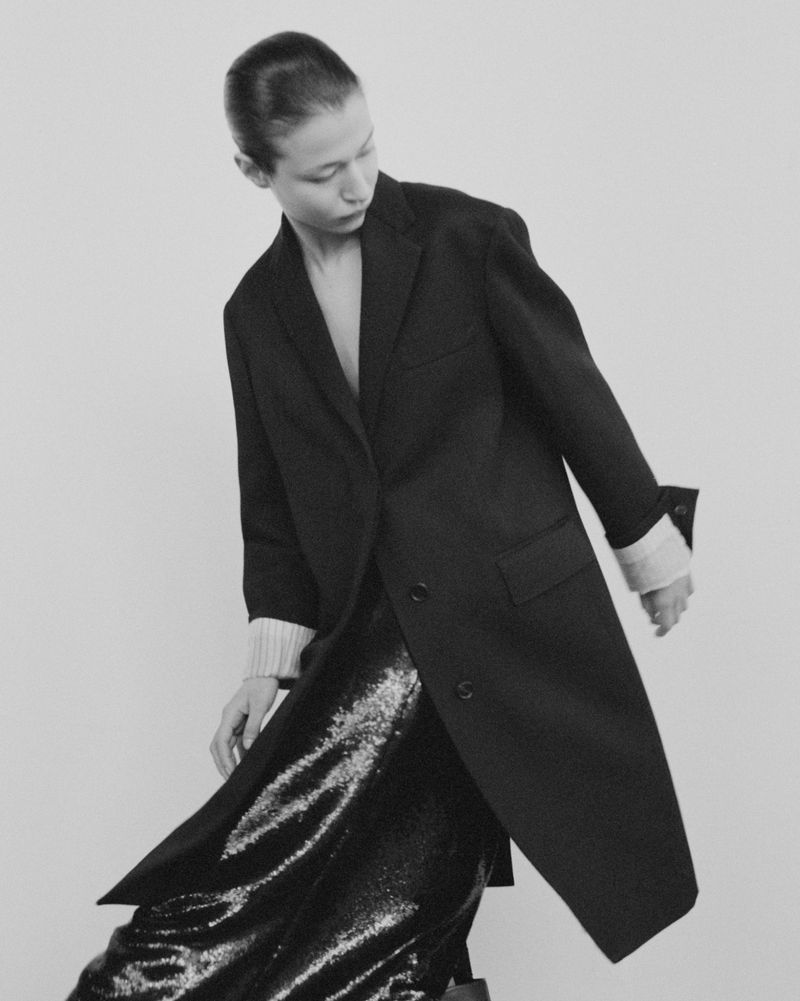 Monochrome fashion shot of a young adult woman in a dark coat over a sequin skirt, posing against a plain background in a studio.