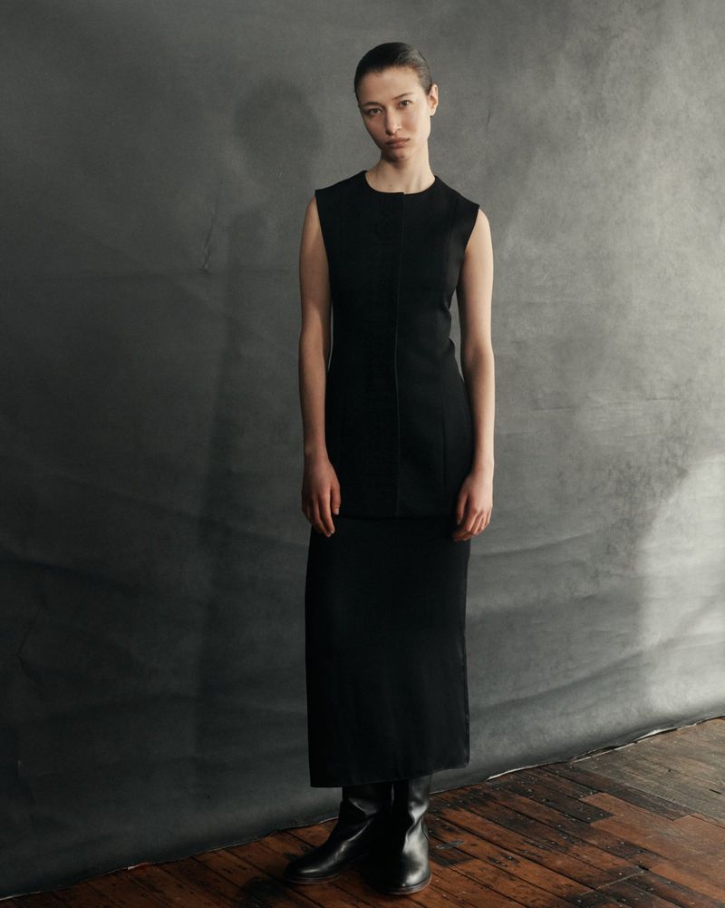 Leyla Greiss stands wearing a sleeveless black tunic, matching long skirt, and black leather boots against a grey studio backdrop.
