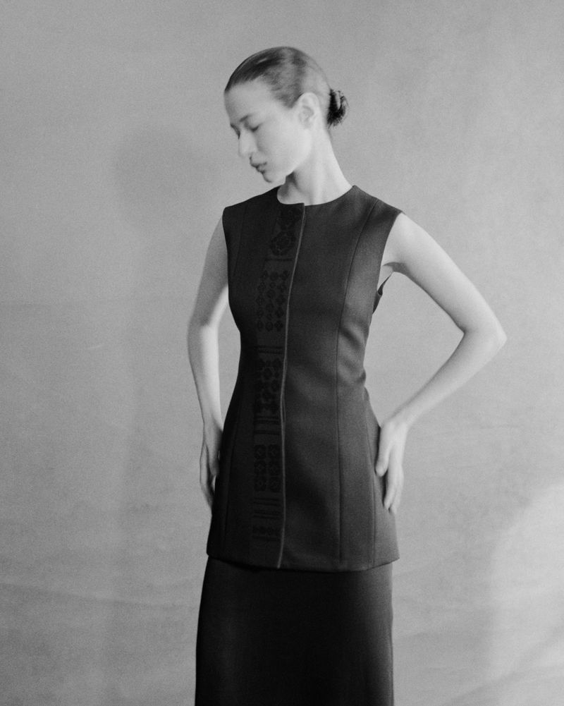 Monochrome portrait of a woman in a dark sleeveless top and skirt, hands on hips, looking down in a neutral indoor setting.