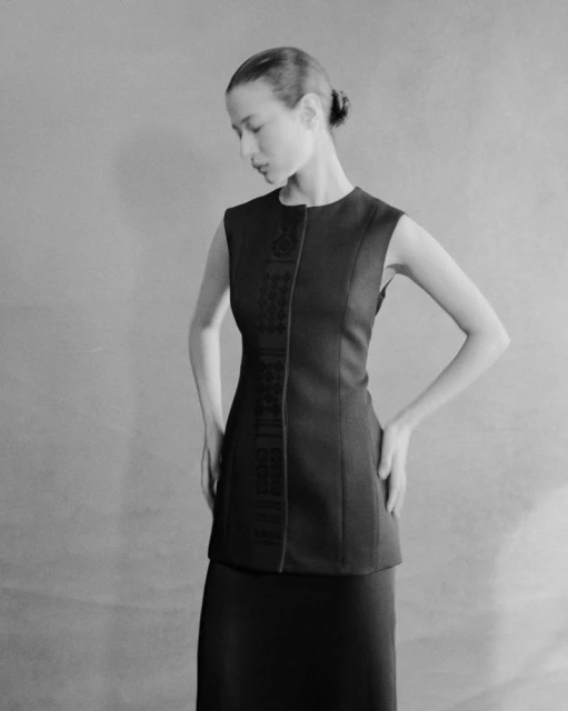 Monochrome portrait of a woman in a dark sleeveless top and skirt, hands on hips, looking down in a neutral indoor setting.