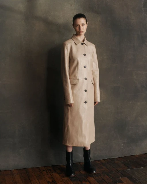 Woman in a tan buttoned coat and black boots poses in front of a mottled gray background with a hardwood floor in a fashion editorial.