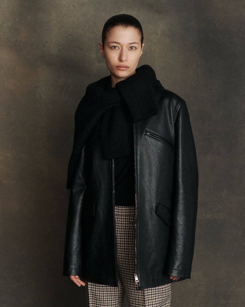 Adult woman in black leather jacket and plaid pants poses against a brown background, creating a high-fashion editorial portrait.