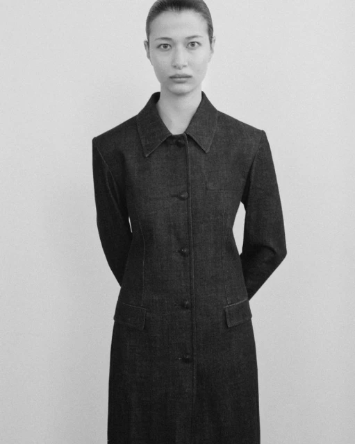 Black-and-white studio shot of a slender adult woman in a dark, buttoned denim dress with hands clasped behind her in neutral pose.