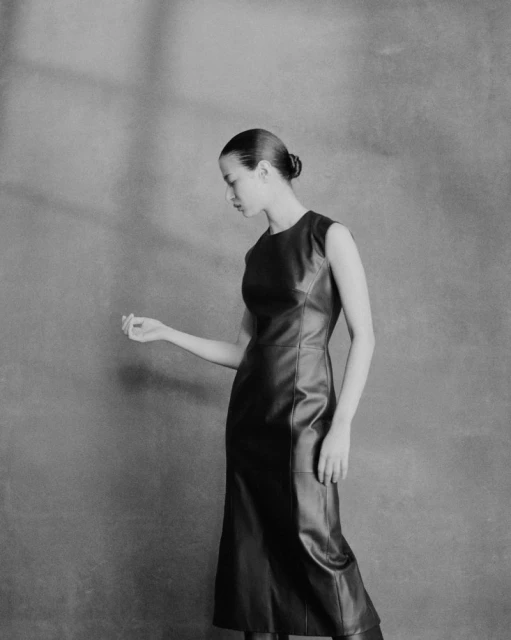 A woman poses in a black sleeveless leather dress, looking down in a studio setting with soft lighting and a neutral background.