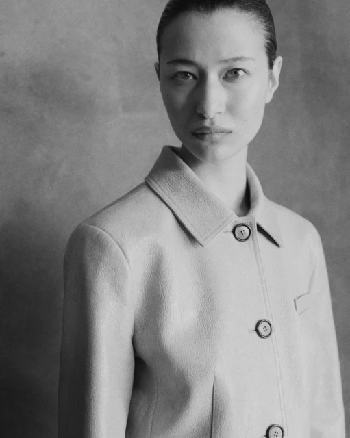 Monochrome portrait of an adult woman in a light-colored jacket with buttons, looking directly at the camera against a plain backdrop.