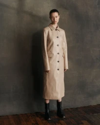 Model Leyla Greiss standing in a long beige button-front leather coat and black boots against a textured dark grey backdrop.