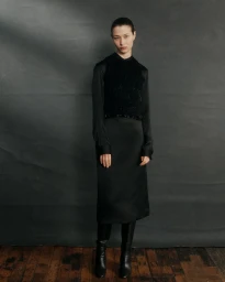 Model Leyla Greiss wearing a black satin midi dress, textured knit vest, and knee-high leather boots against a dark grey backdrop.