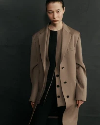 Model Leyla Greiss wearing a layered taupe tailored coat over a black top against a dark studio backdrop.