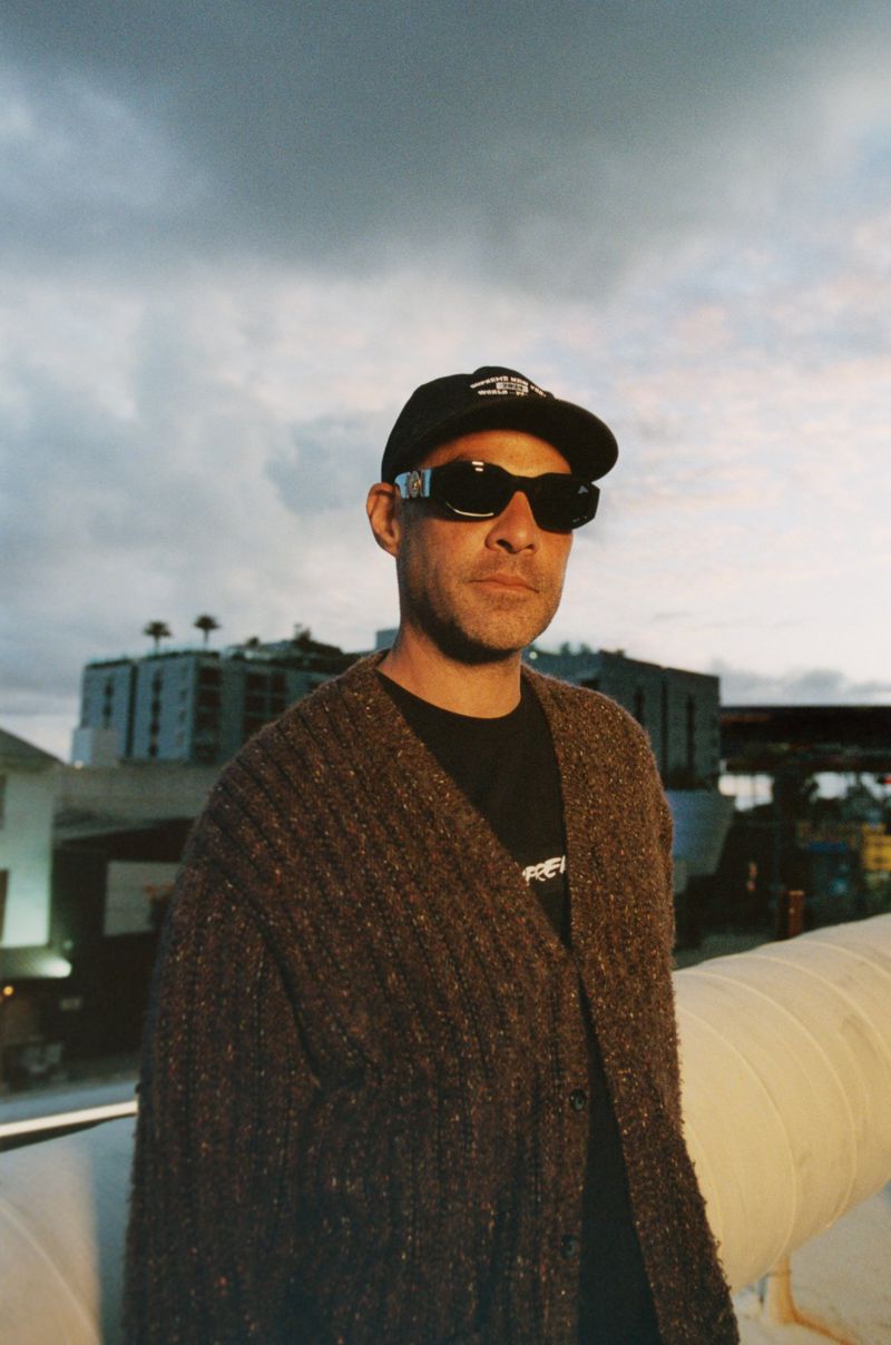 Portrait of a man wearing a black t-shirt, cardigan, hat, and sunglasses, standing on a rooftop with a city skyline and cloudy sky.