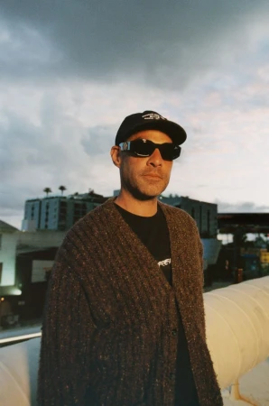 Portrait of a man wearing a black t-shirt, cardigan, hat, and sunglasses, standing on a rooftop with a city skyline and cloudy sky.