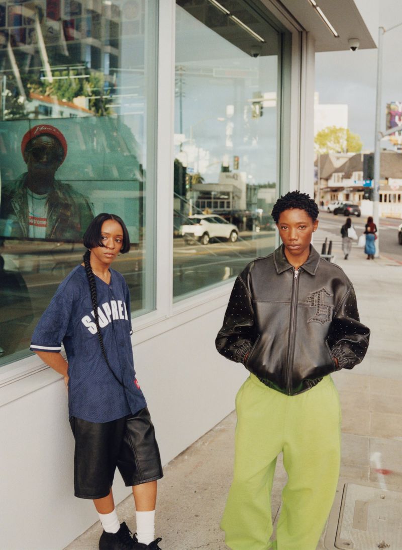 Two adult women pose outside a building with large windows; one wears a baseball shirt, and the other, a leather jacket and green pants.