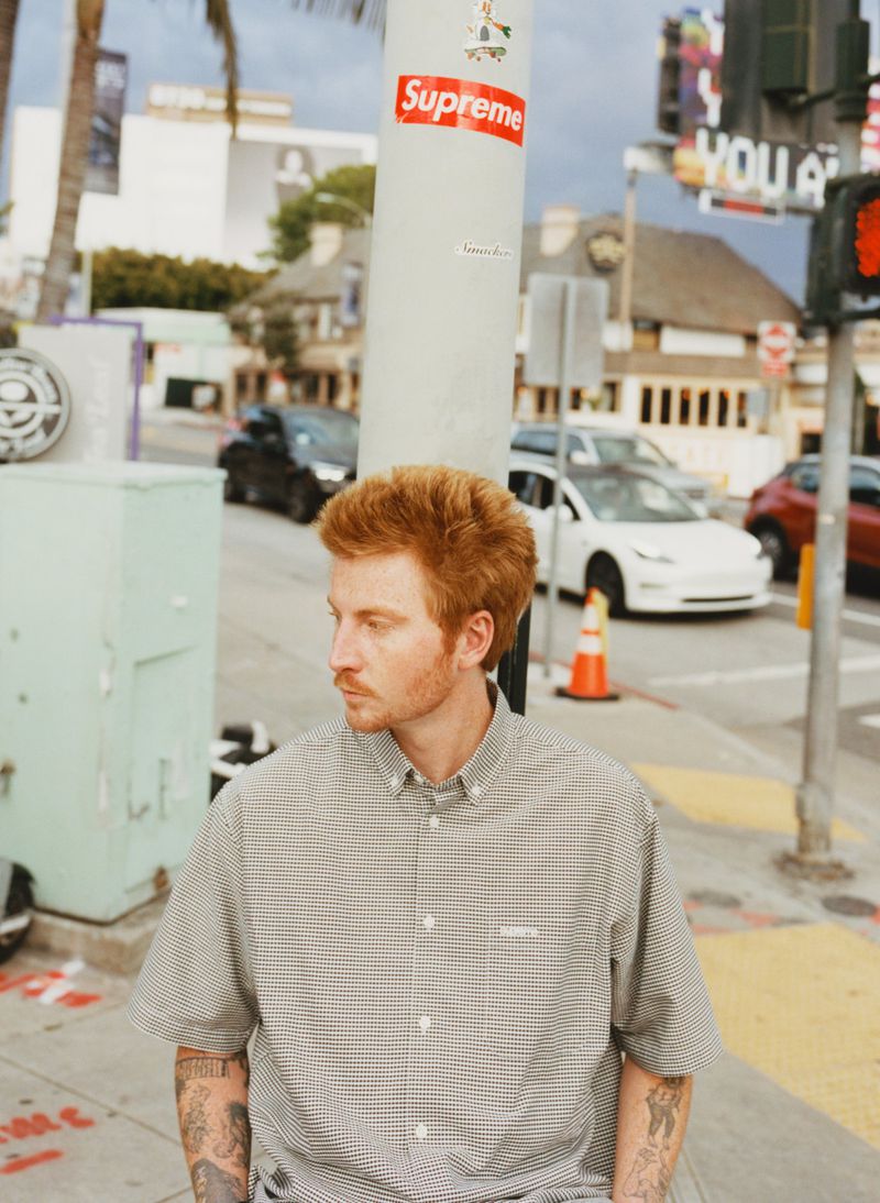 Red-haired man in patterned shirt stands on a sidewalk in front of city street scene on a sunny day with sky and palm trees.