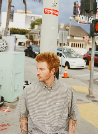 Red-haired man in patterned shirt stands on a sidewalk in front of city street scene on a sunny day with sky and palm trees.