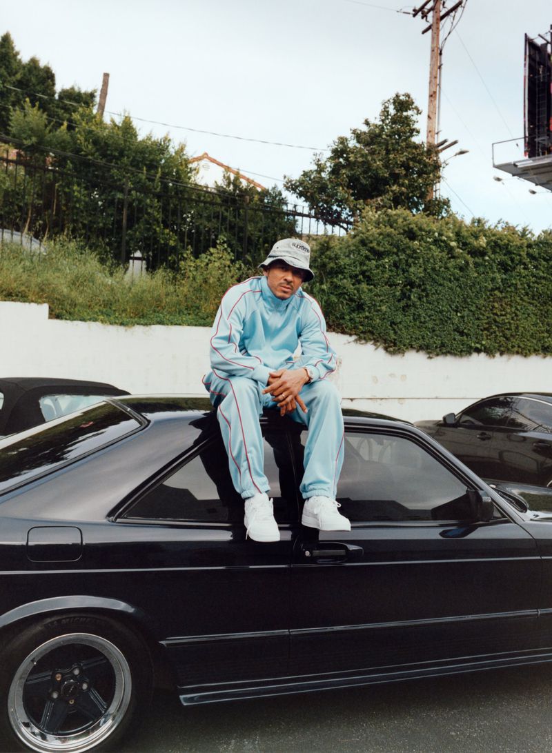 Man in a blue tracksuit and bucket hat sitting on the hood of a black car in an outdoor setting with lush greenery.