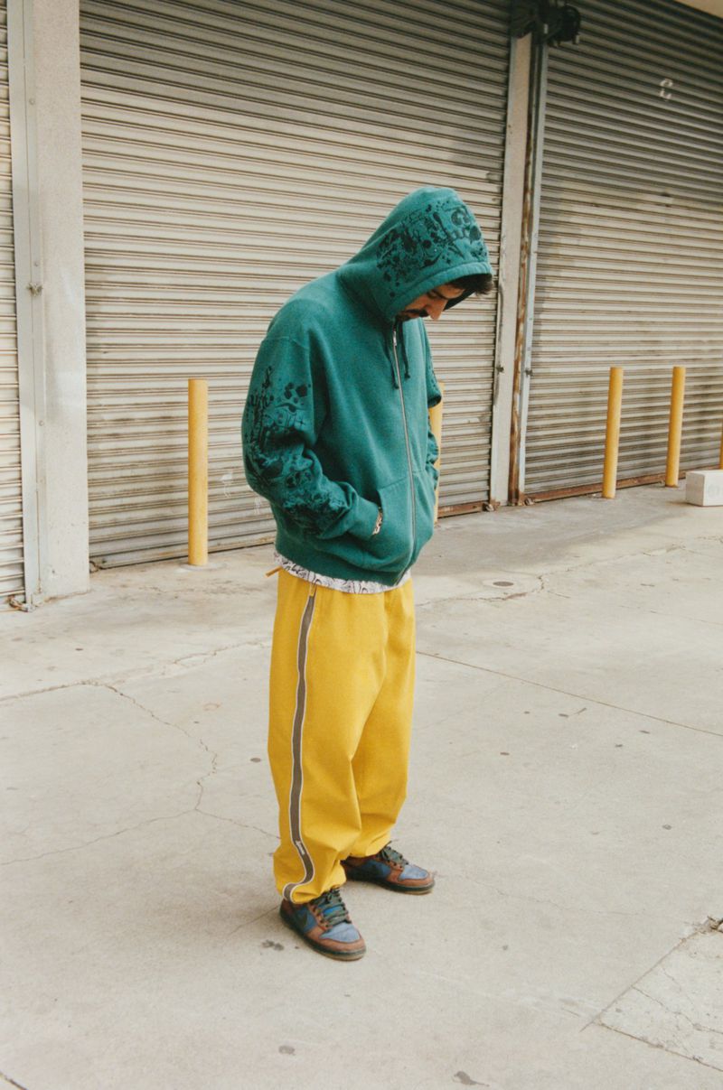 Person in green hoodie and yellow pants stands near gray metal shutters and yellow bollards on concrete in daytime outdoor shot.