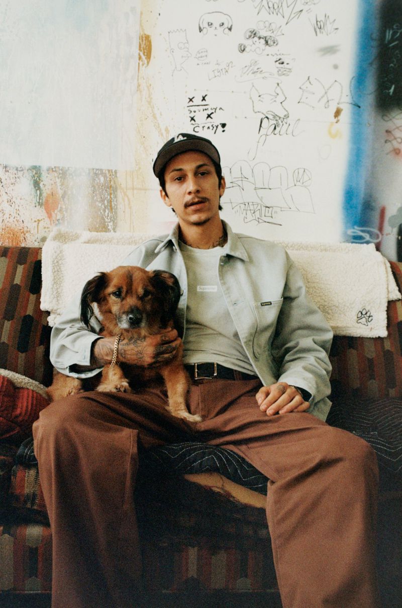 Young adult male poses on an patterned couch, cradling a brown dog. The backdrop features wall art and light indoor lighting.