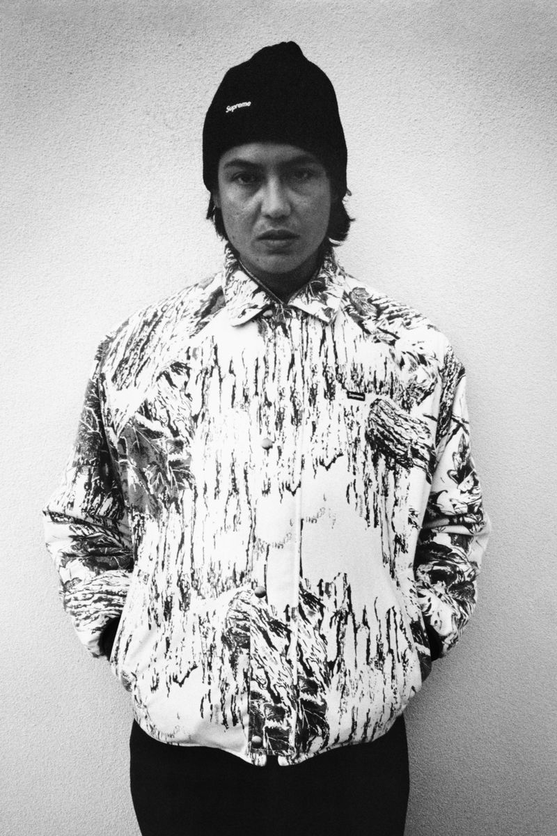 Monochrome portrait of an adult wearing a patterned jacket and beanie hat against a grey backdrop in an indoor setting.