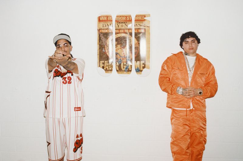 Two adult males stand indoors against a white wall, one wearing a jersey and shorts and the other in an orange tracksuit. 