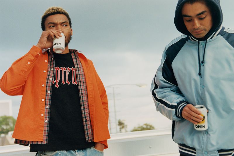Two young men outdoors: one drinking from a white can in an orange jacket, the other in a blue jacket holding a can.