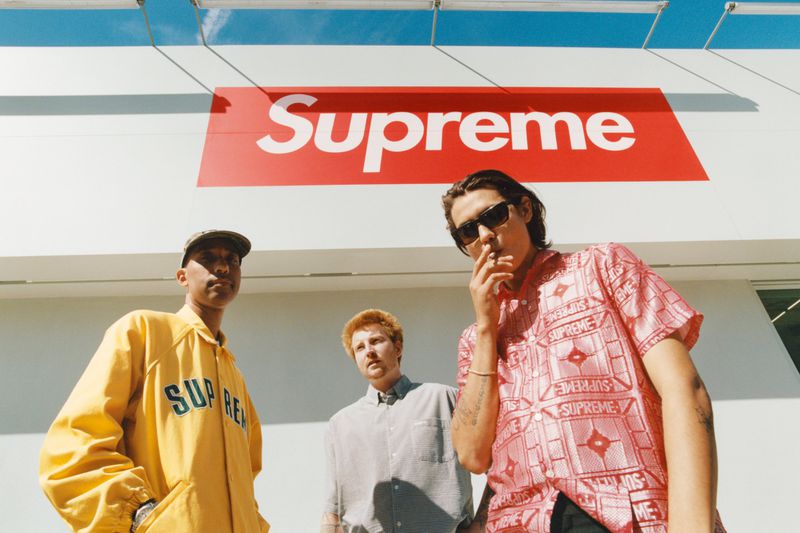 Three men stand outside with a 'Supreme' sign in the background. One wears sunglasses and smokes a cigarette.