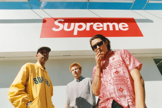 Three men stand outside with a 'Supreme' sign in the background. One wears sunglasses and smokes a cigarette.