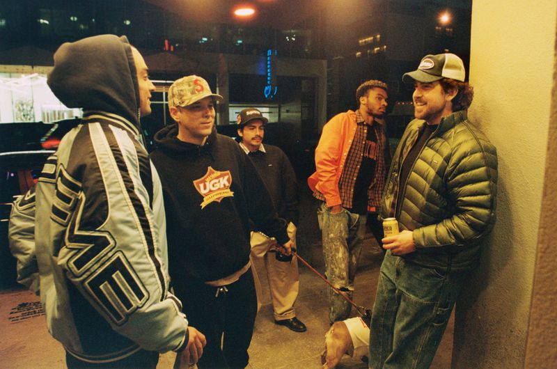 Group of five casually dressed people and a dog gather outside an urban building at night with neon lighting in the background.