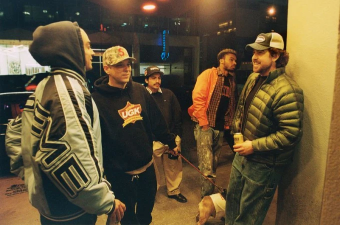 Group of five casually dressed people and a dog gather outside an urban building at night with neon lighting in the background.