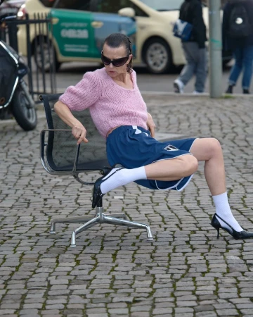 Adult woman in pink sweater and blue shorts sitting on black office chair in urban outdoor setting with cars in the background.