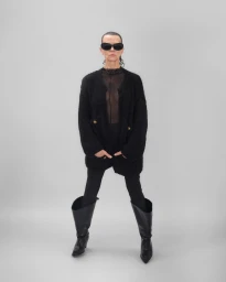 Mature woman in black sunglasses, a textured black cardigan, slim trousers, and wide black knee-high boots on a grey backdrop.