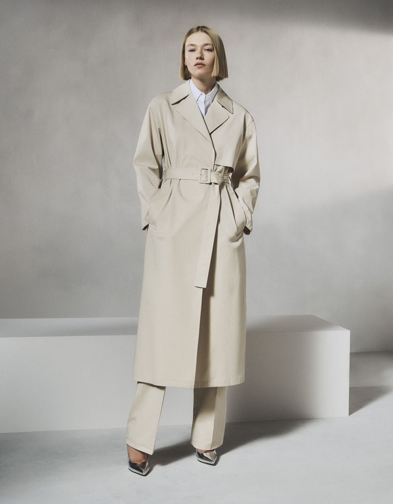 Woman poses in a beige trench coat and matching pants. She is standing indoors, and a neutral background and white cube are visible.