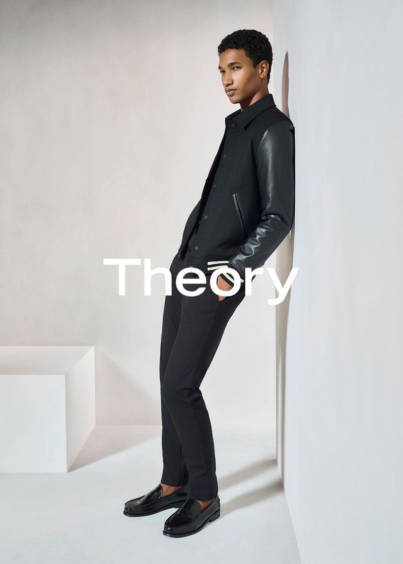 Fashion shot of a young adult man in a black jacket and trousers leaning against a white wall in a studio, clean background.