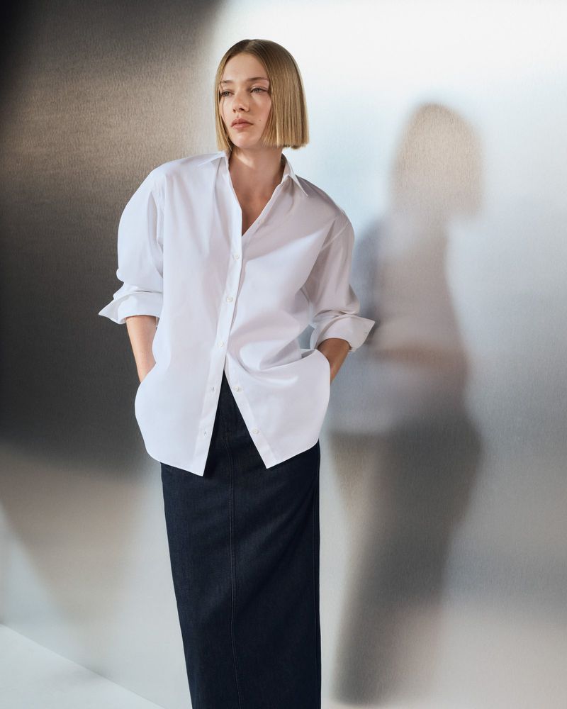 Model Ella Rattigan wearing an oversized white button-up shirt and dark denim maxi skirt against a metallic studio backdrop.