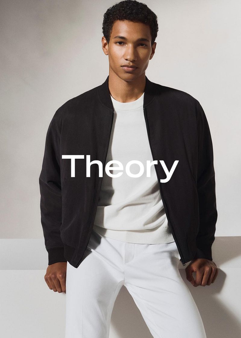 A fashionable black male model is posing indoors wearing a Theory jacket, white t-shirt and matching pants for a studio shot.