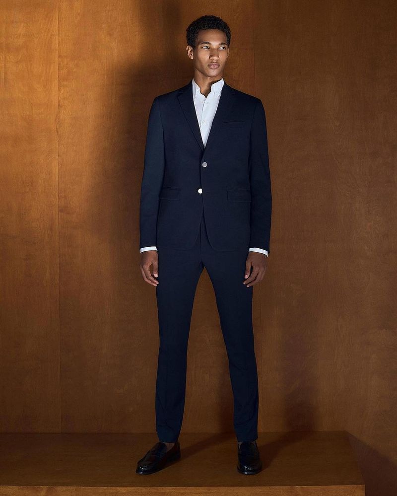 A young, dark-haired male model poses in a navy suit with a white shirt and black loafers against a warm brown wood background.