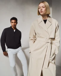 Model in a beige belted trench coat posed in front of a man in a black jacket and white trousers against a neutral studio backdrop.