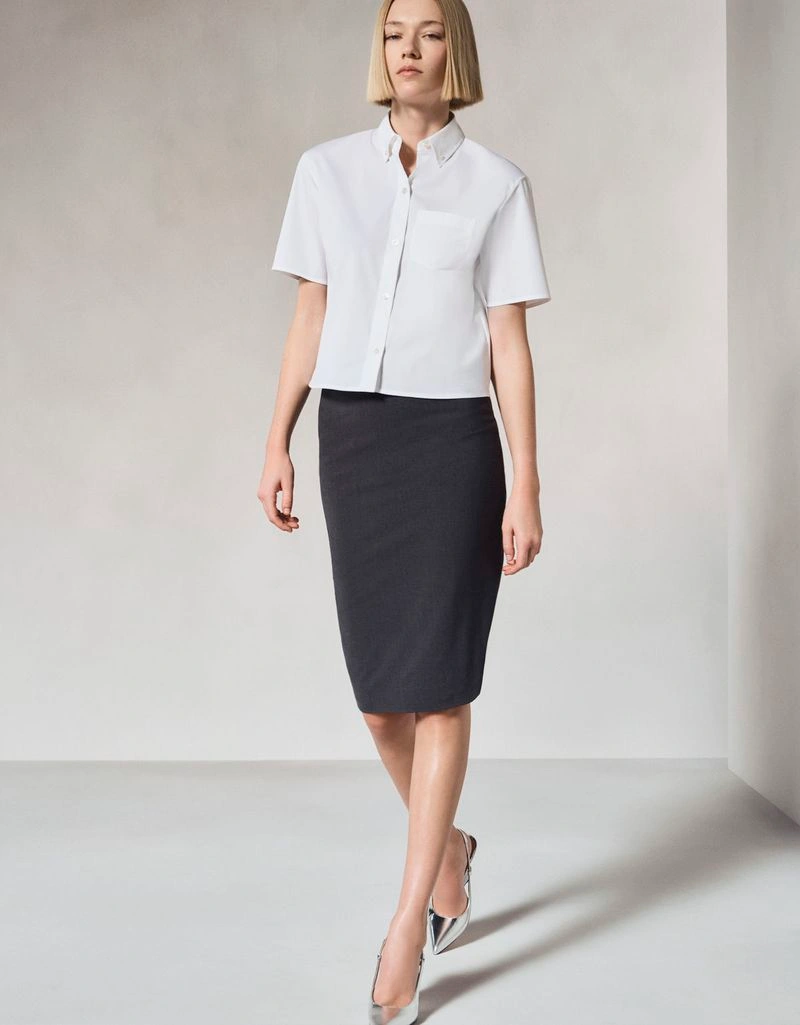 Model Ella Rattigan in a white short-sleeve button-down shirt and black pencil skirt walking in a minimalist studio.