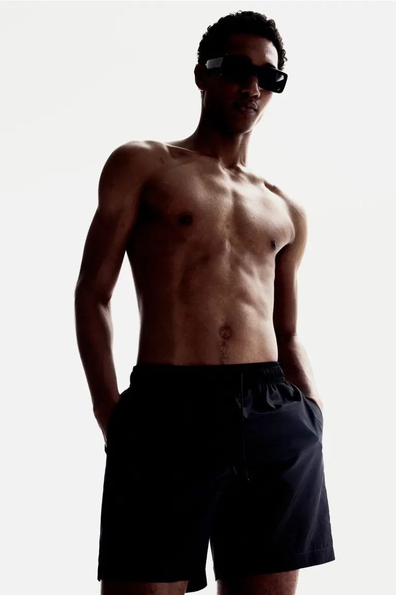 An adult male model posing shirtless, wearing dark swim shorts and sunglasses, against a bright white background in a studio setting.