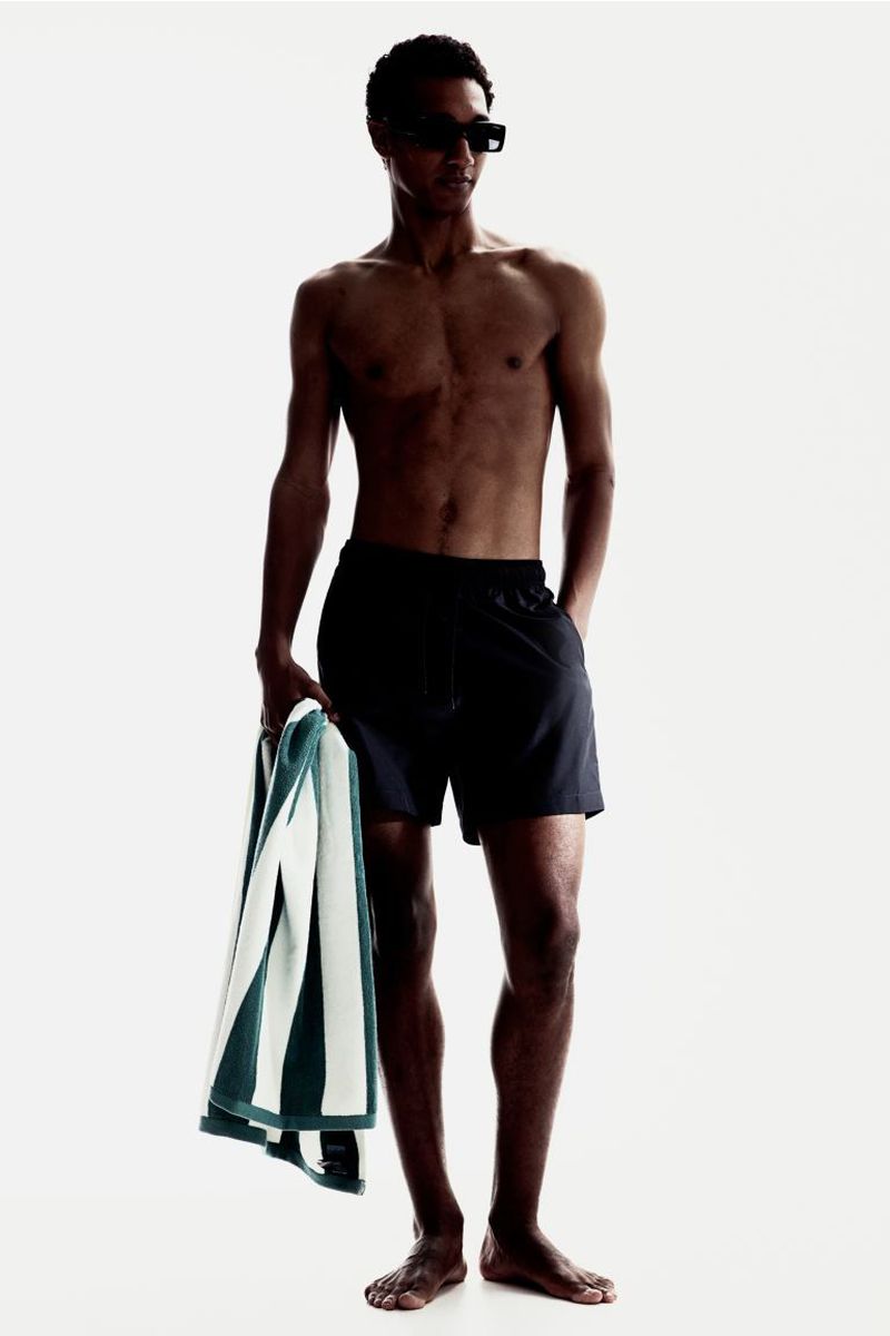 Joshua Seth standing shirtless in black swim trunks and sunglasses while holding a striped green towel against a white background.