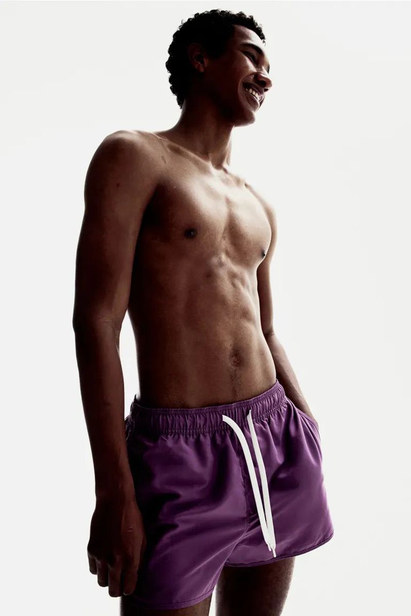 Athletic man in purple swim trunks smiles in a brightly lit, high-key studio portrait, showcasing swimwear fashion.