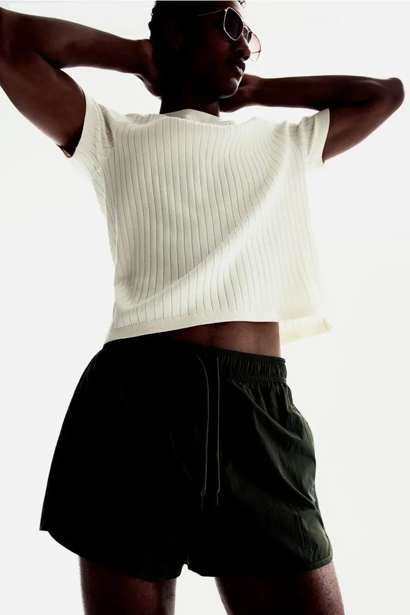 Athletic African-American man in sunglasses poses in a studio wearing a ribbed white t-shirt and dark olive-green running shorts.