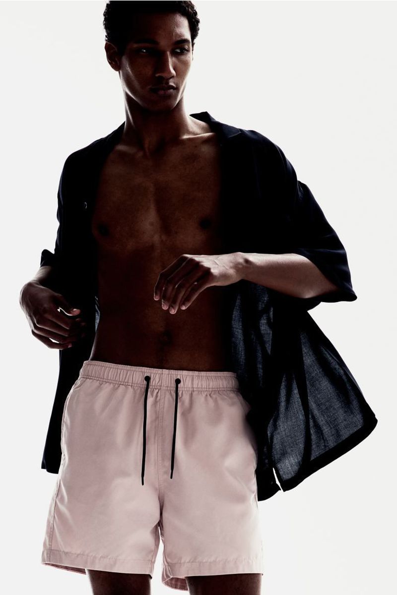 Adult male poses in pink shorts and open black shirt against a white background in a fashion style portrait, indoors with studio lighting.