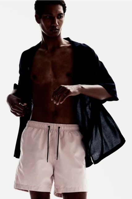 Adult male poses in pink shorts and open black shirt against a white background in a fashion style portrait, indoors with studio lighting.