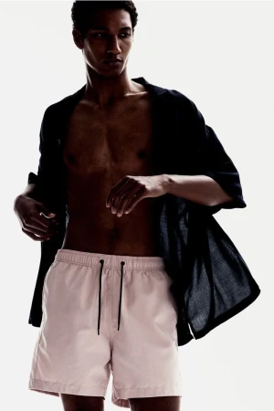Adult male poses in pink shorts and open black shirt against a white background in a fashion style portrait, indoors with studio lighting.
