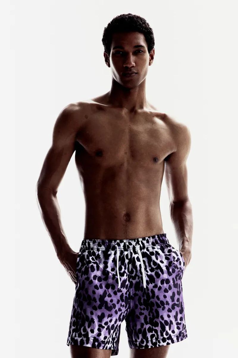 A dark-skinned, muscular adult male poses in a bright studio wearing animal-print swim trunks with his hands in his pockets.
