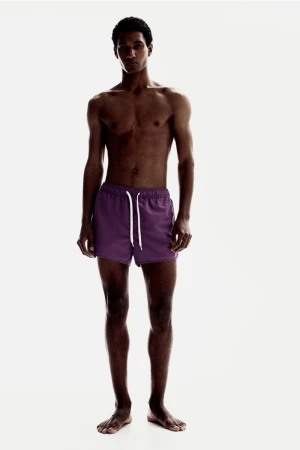 Shirtless male model Joshua Seth wearing purple swim trunks with a white drawstring against a plain white studio backdrop.