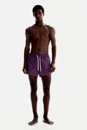 Shirtless male model Joshua Seth wearing purple swim trunks with a white drawstring against a plain white studio backdrop.
