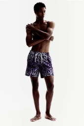 Model Joshua Seth posing shirtless in purple leopard print swim shorts against a seamless white studio background for H&M.