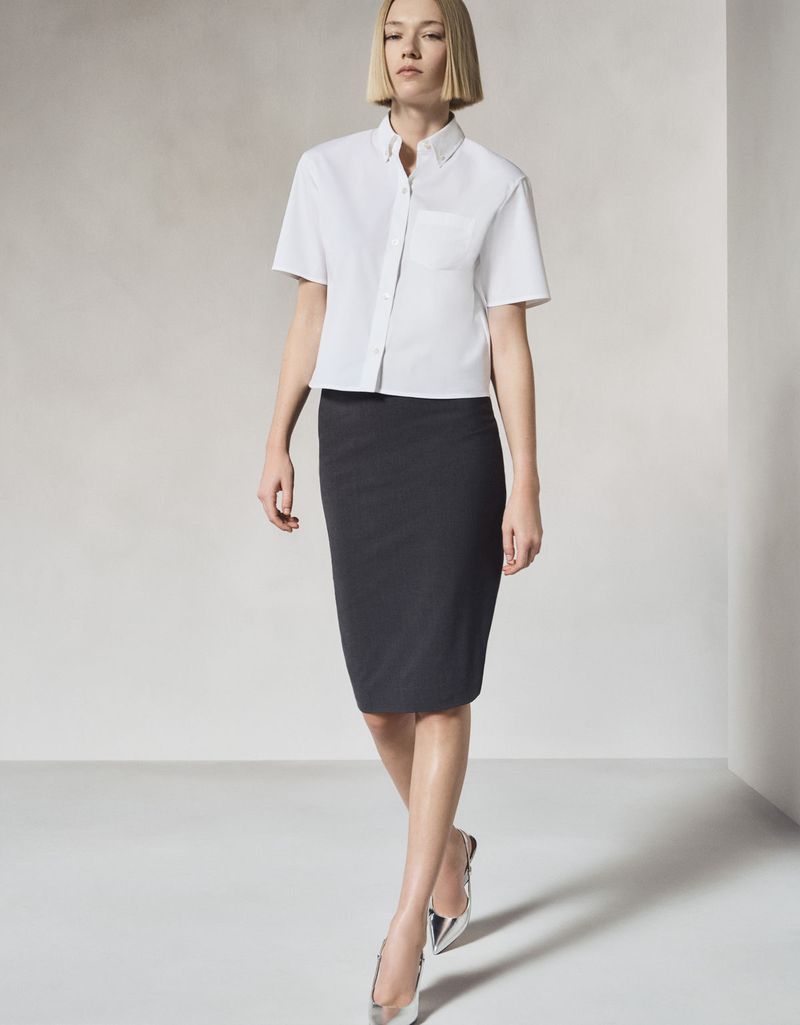 Full-shot of a blond-haired adult woman in a white short-sleeved shirt, pencil skirt, and silver flats against a neutral background.